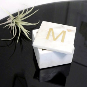 May include: White marble box with a gold letter "M" on the lid. The box is open and sitting on a black surface.