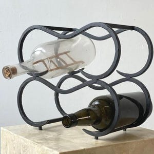 May include: Black wrought iron wine rack holding two bottles. One bottle is clear glass with a cork, the other is green glass. The rack has a decorative, looping design and is displayed on a light-colored surface.
