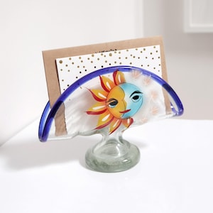 Peut inclure: Un porte-cartes en verre avec un bord bleu et un motif peint de soleil et de lune. Le porte-cartes a une base en verre transparent.