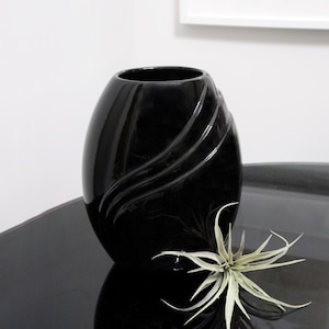 May include: A black ceramic vase with a wavy design. The vase is sitting on a black surface with a small green plant in front of it.