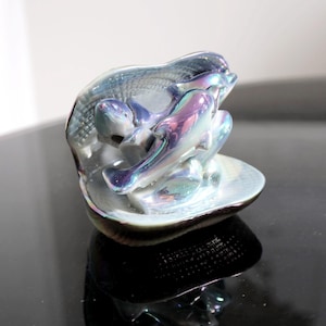 May include: A iridescent ceramic figurine of two dolphins jumping out of a seashell. The figurine has a pearlescent finish and is a decorative piece.