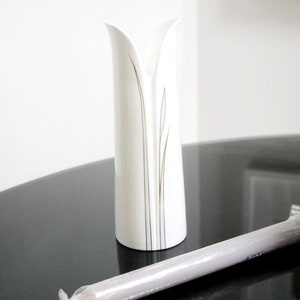 May include: A white ceramic vase with a gold and blue design. The vase has a narrow opening and a wide base. It is sitting on a black surface.