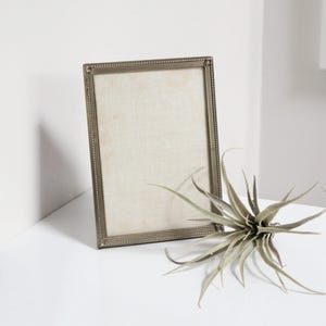 Peut inclure: Un cadre photo argenté avec une bordure perlée et un passe-partout blanc. Le cadre est posé sur une surface blanche avec une petite plante verte au premier plan.