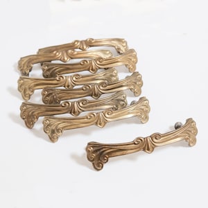 May include: A collection of antique brass drawer pulls with an ornate, scroll-like design. The hardware is a warm, golden color and features a curved shape with decorative detailing. These vintage pulls are ideal for furniture restoration projects.