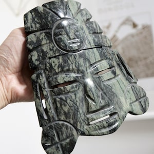 May include: A green stone mask with intricate carvings of a face and a sun design. The mask is made of a dark green stone with white veins running through it.