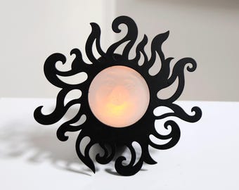 May include: A black, sun-shaped decorative light fixture with a glowing, round center. The sun's rays are stylized with swirling, curved edges. The light fixture is placed on a white surface.