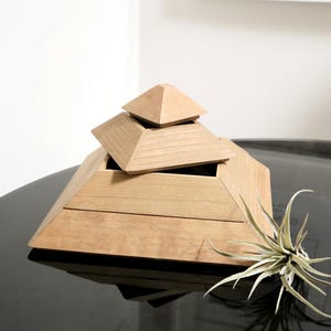 May include: A wooden pyramid-shaped box with a tiered design. The box is made of light-colored wood and has a removable lid. A small air plant sits to the side of the box. The box is on a black surface.