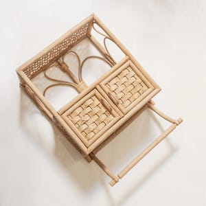 May include: A natural woven rattan storage shelf with two small cabinet doors and a towel bar. The shelf has a rectangular shape with a woven top and decorative curved accents. The cabinet doors have a woven pattern.