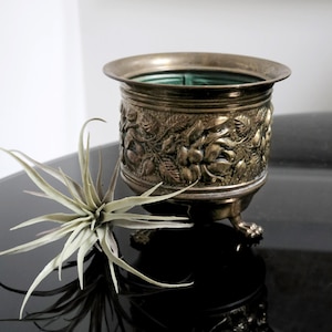 Könnte beinhalten: Ein goldfarbener Metalltopf mit einem verzierten Blumenmuster und drei Füßen. Der Topf steht auf einer schwarzen Oberfläche.