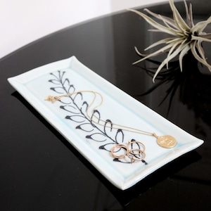 May include: A light blue ceramic tray with a black floral design. A gold necklace and earrings are on the tray.