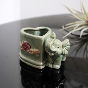 May include: A green ceramic planter shaped like a heart with an elephant figurine attached to the side. The planter has a floral design and the elephant has a smiling face.