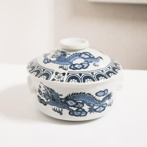 May include: A white ceramic lidded bowl with blue dragon and cloud designs. The bowl has two small handles and a small knob on the lid. The rim of the bowl and lid feature a repeating pattern of arches and floral elements.