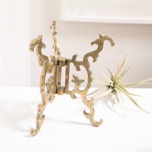 May include: Ornate brass-colored metal stand with a decorative, openwork design. The stand has a three-legged base and a central support structure. The stand is designed to hold a small object. A plant is in the background.