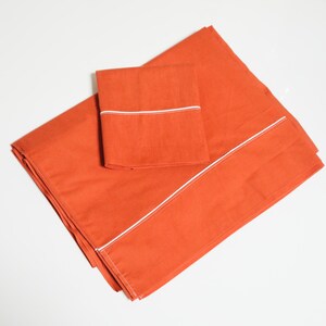 May include: An orange bed linen set, comprising a flat sheet, fitted sheet, and pillowcase. The sheets feature a white trim. The bedding is neatly folded and arranged on a white surface.