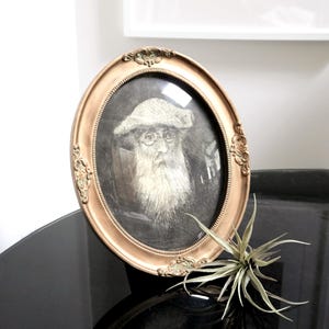 May include: An oval-shaped gold picture frame with a black and white portrait of a man with a beard and a hat. The frame has ornate details and is sitting on a black surface.