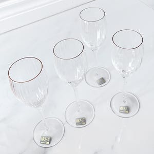 Può includere: Quattro calici da vino in vetro trasparente con texture a coste verticali sui bevanti e steli sottili. Ogni calice presenta un bordo dorato e un piccolo adesivo rettangolare sulla base con la scritta Mikasa.