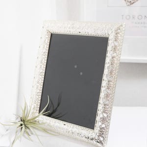 May include: A rectangular picture frame with a metallic silver finish and an intricate, embossed floral filigree pattern along the border. The frame features a smooth, black interior panel and a beaded inner edge detail surrounding the center.
