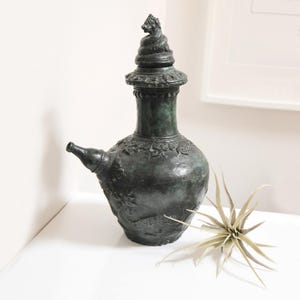 May include: An antique-style, dark green water vessel with a spout and decorative detailing. The vessel features a lid with a sculpted animal figure. A dried plant with spiky leaves is placed nearby.
