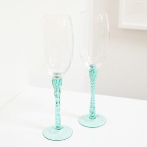 May include: Two clear champagne flutes with a twisted, seafoam green stem and base. The glasses are empty and sit on a white surface. The stems have a decorative, rope-like design.