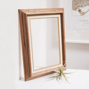 May include: A rectangular wooden picture frame with a light beige mat. The frame has a natural wood grain finish. A small air plant with green and silver-gray leaves rests near the frame. The frame is leaning against a white wall.