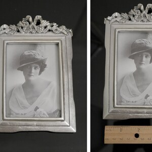Pu&ograve; includere: Una cornice argentata con un designo ornato e una foto in bianco e nero di una donna con un cappello. La foto &egrave; intitolata "Adele, 18 anni."