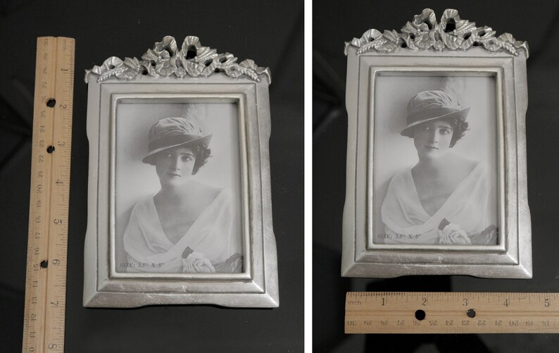 Pu&ograve; includere: Una cornice argentata con un designo ornato e una foto in bianco e nero di una donna con un cappello. La foto &egrave; intitolata "Adele, 18 anni."