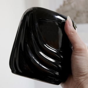 May include: A black ceramic planter with a wavy, organic design. The planter is being held in a hand.