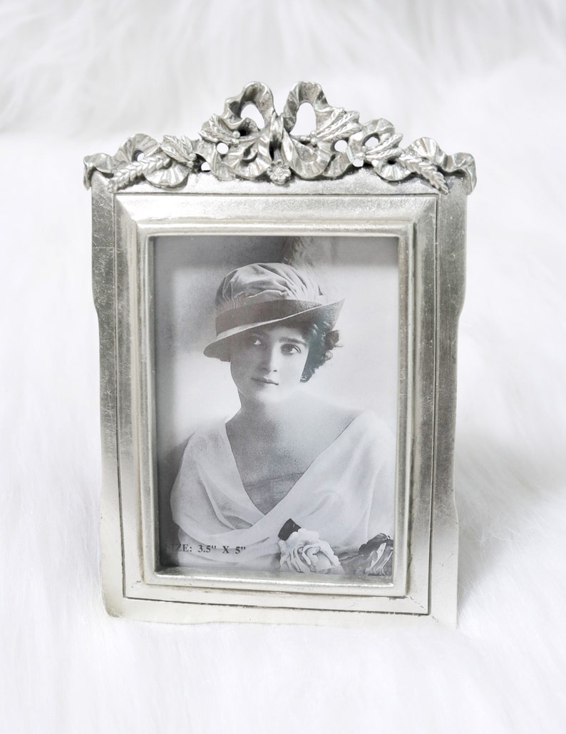 Pu&ograve; includere: Una cornice per foto argentata con un designo ornato, che presenta un fiocco e un motivo a foglie in rilievo. La cornice contiene una fotografia in bianco e nero di una donna con un cappello e un vestito bianco. La foto &egrave; sottotitolata "Size: 3.5" x 5""