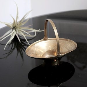 May include: A small, gold-colored, decorative bowl with a handle. The bowl has an ornate design and is sitting on a black surface.