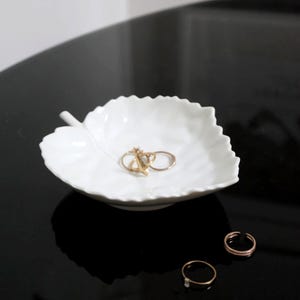 Könnte beinhalten: Weiße, blattförmige Schale aus Keramik mit goldenen Ringen und einem goldenen Stab im Inneren. Die Schale steht auf einer schwarzen Oberfläche.