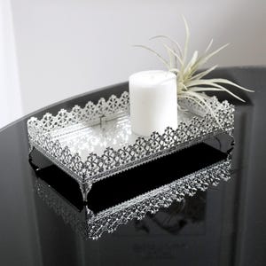 May include: A silver decorative tray with a delicate filigree design. The tray has a mirrored surface and is sitting on a black surface. A white candle and a small plant are on the tray.