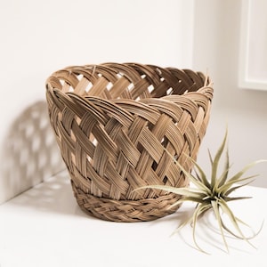 May include: A light brown woven basket with a flared top and a round base. The basket has a crisscross pattern. A green plant with long, thin leaves sits beside the basket on a white surface.
