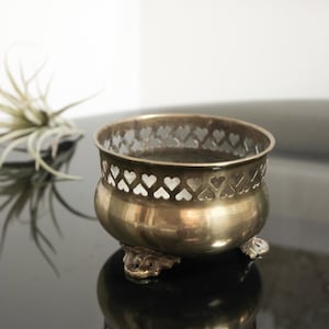 May include: A small, antique brass bowl with a heart-shaped cut-out design around the rim. The bowl sits on three ornate feet. The bowl is reflective and sits on a dark surface. A plant is in the background.