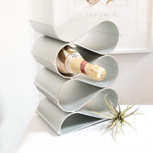 May include: A gray metal wine rack with a wave-like design, holding a bottle of champagne. The rack has four curved sections to cradle the bottles. The champagne bottle has a gold label and a red and gold foil top.
