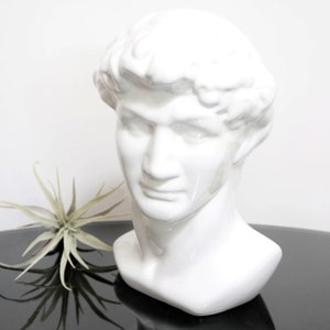May include: White ceramic bust sculpture of a man's head and neck. The bust is smooth and has a classic style.