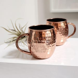 May include: Two hammered copper mugs with handles. The mugs have a shiny, metallic finish.