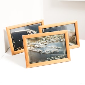 May include: Three framed landscape photographs with wooden frames. The photographs depict coastal scenes, with one labelled "Clarks Harbour Nova Scotia" and another "Port La Nova Scotia". The frames are light brown.