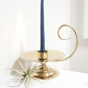 May include: A brass candlestick holder with a navy blue candle. The holder has a circular base and a curved handle. An air plant is placed next to the candlestick holder. The candle holder is a decorative home decor item.