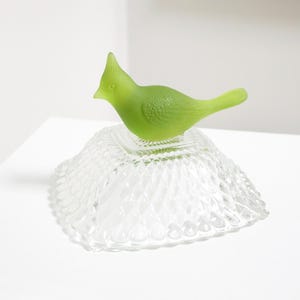 May include: A green glass bird ornament sits on a clear, diamond-patterned glass dish. The bird has a crest and a textured body. The dish is square with a scalloped edge. Decorative home decor.