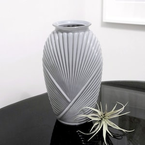 May include: A grey ceramic vase with a fan-shaped design. The vase is oval-shaped and has a smooth, glossy finish.