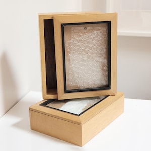 Könnte beinhalten: Zwei Holzkisten mit Glasplatten und schwarzen Rändern. Eine Kiste ist geöffnet und gibt den Innenraum frei. Die andere Kiste ist geschlossen. Die Kisten sind aus hellem Holz und stehen auf einer weißen Oberfläche.