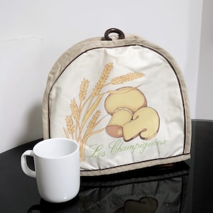 May include: A beige and brown fabric toaster cover with a mushroom and wheat design. The text "Les Champignons" is printed on the cover.