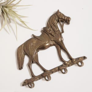 May include: A bronze-coloured horse-shaped wall hook with a saddle and four hooks below. The horse is in a rocking horse pose, with a detailed mane and tail. The hook is made of metal.