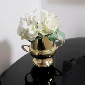 Könnte beinhalten: Eine goldfarbene Metallvase mit rundem Sockel und zwei Henkeln, gefüllt mit weißen Hortensien und Rosen. Die Vase steht auf einer schwarzen, reflektierenden Oberfläche, was einen Kontrast erzeugt.