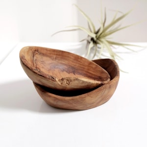 May include: Two wooden bowls, one nested inside the other, are made of natural wood with a light brown colour. The bowls have a rounded, oval shape and a smooth, slightly textured surface.