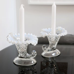 May include: Pair of clear glass candle holders with a white opalescent finish. The holders are shaped like a cornucopia with a ruffled edge and a beaded texture. Each holder has a white candle.