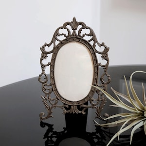 May include: An ornate, oval-shaped picture frame made of dark brown metal with intricate scrollwork details. The frame has a stand and is sitting on a black surface.