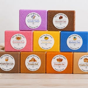 May include: A pyramid of nine rectangular bars of handmade vegan soap in various colors, each featuring a circular label with the brand name Angie's Soap Bakery Company, a scent name like Lavender Vanilla or Pumpkin Pie, and a 5oz weight label.