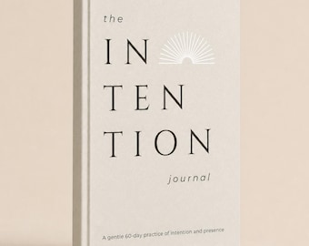 May include: A light beige hardcover journal with the title "the INTENTION journal" printed in dark brown text on the front cover. A white sunburst graphic is positioned above the word "INTENTION," and the subtitle reads "A gentle 60-day practice of intention and presence."