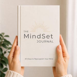 May include: A white hardcover journal titled The MindSet Journal with a gold sunburst graphic above the text and the subtitle 40 Days to Reprogram Your Mind printed in black. The cover features a clean design with centered typography and lines.